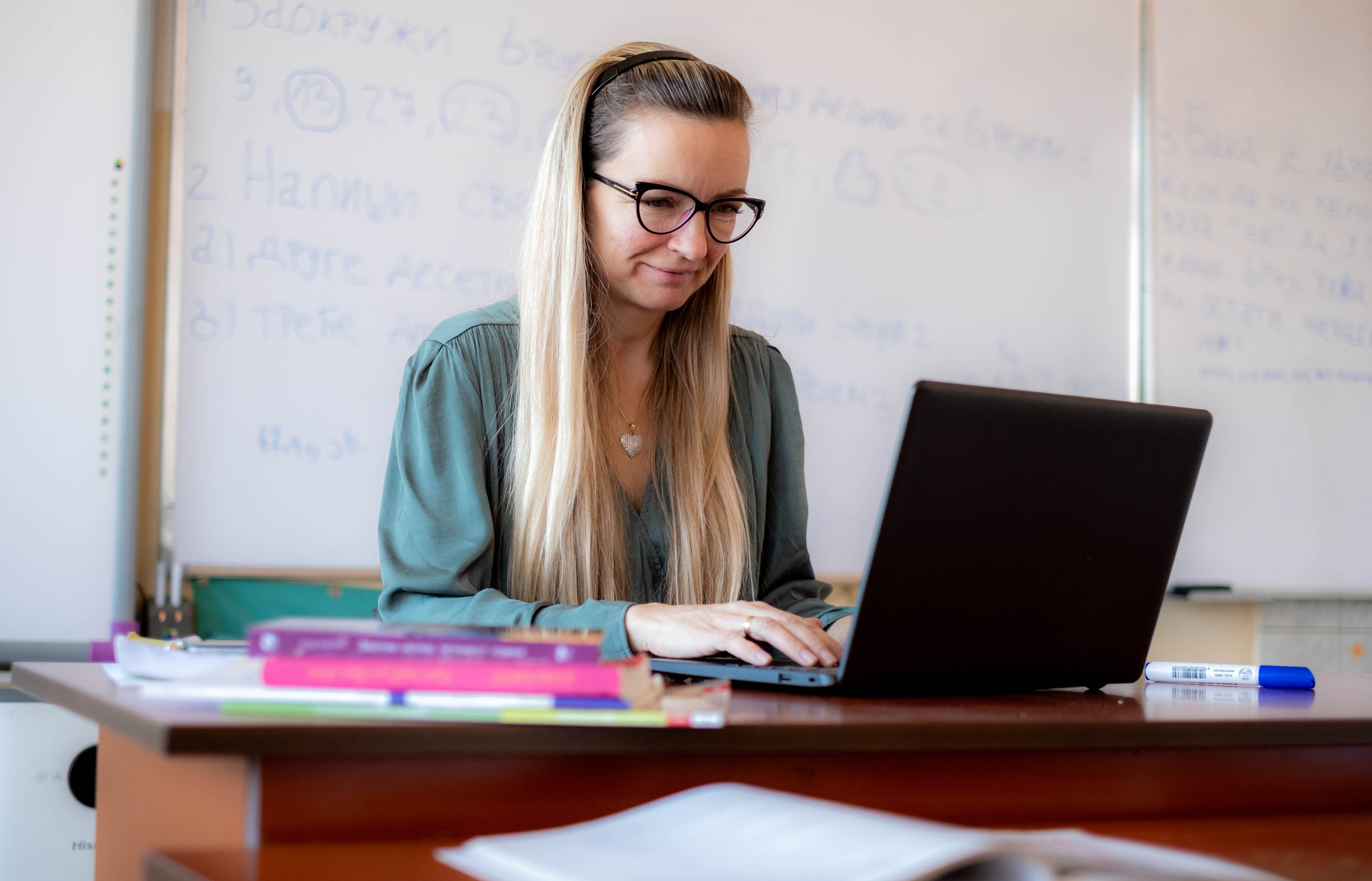 A teacher is sitting at her desk and working on her laptop.
